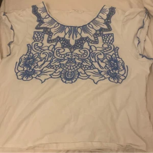 Vit t-shirt med blått blommönster - Vit t-shirt med korta ärmar och ett stort, detaljerat blått blommönster framtill. Rund halsringning och mjukt material som känns skönt mot huden. Perfekt för dig som gillar unika prints och vill sticka ut lite extra.