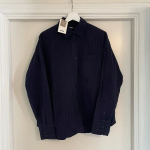 Mörkblå randig skjorta från BikBok - Snygg mörkblå skjorta från BikBok med tunna vita ränder. Klassisk krage, knäppning framtill och en bröstficka. Skjortan är i bomull och har långa ärmar med manschettknappar. Perfekt för en clean och stilren look.