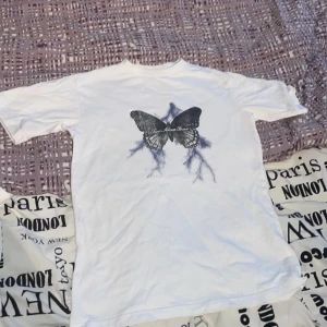 Vit t-shirt med fjärilstryck - Snygg vit t-shirt med ett stort fjärilstryck i svart och blått på framsidan. Perfekt för en avslappnad stil med en unik touch. Passar bra till jeans eller shorts.