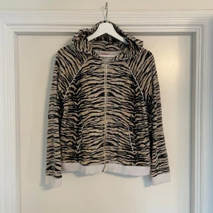Zebramönstrad hoodie Juicy Couture - Säljer en beige och svart hoodie från Juicy Couture med zebramönster. Tröjan har dragkedja framtill, huva och långa ärmar. Mjukt material som känns skönt mot huden, perfekt för chill dagar. Fickor på sidorna och ribbade muddar vid ärmslut och nederkant.