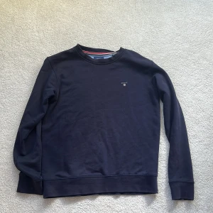 Mörkblå sweatshirt från GANT - Klassisk mörkblå sweatshirt från GANT med rund halsringning och diskret logga broderad på bröstet. Tröjan har långa ärmar och ribbade muddar vid ärmslut och nederkant. Perfekt för en clean och stilren look.
