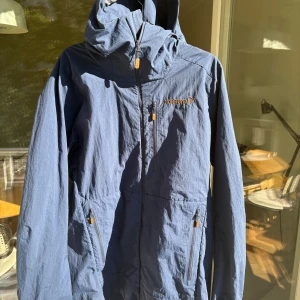Blå vindjacka från Norrøna, M - Snygg blå vindjacka från Norrøna i modellen Svalbard Lightweight. Jackan har huva, dragkedjor med orange detaljer och flera fickor. Tillverkad i polyester och bomull, perfekt för dig som gillar outdoor och stilren design.