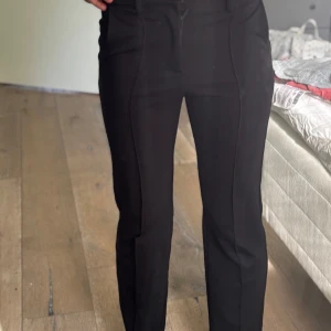 Svarta kostymbyxor från Kappahl - Svarta kostymbyxor från Kappahl i storlek 146. Byxorna har hög midja, raka ben och markerad söm framtill för en clean look. Tillverkade i ett mjukt och stretchigt material som sitter snyggt och bekvämt hela dagen.