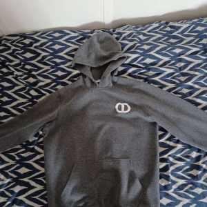Grå hoodie med broderad logga - Snygg grå hoodie med broderad vit logga på bröstet. Klassisk känguruficka framtill och huva. Perfekt för chill dagar eller när du vill ha en enkel men stilren look.