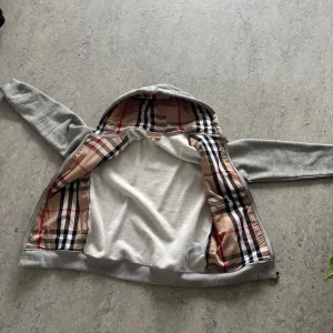 Grå hoodie från Burberry med dragkedja - Säljer en snygg och varm grå Burberry zip-hoodie i storlek M (passar även S/L beroende på passform). Den är ganska ny och endast använd ett fåtal gånger, i mycket fint skick.  ✔️ Klassisk design med dragkedja ✔️ Fickor fram och huva med dragsnören ✔️ Bekväm och stilren  Pris: 1650 kr