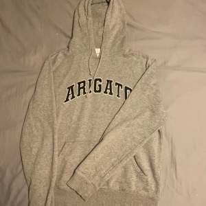 Grå hoodie Axel Arigato - Snygg grå hoodie från Axel Arigato med svartvit logga framtill. Klassisk modell med huva, snörning och känguruficka. Mjuk insida och ribbade muddar. Perfekt för chill dagar eller streetwear-stil.