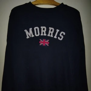 Mörkblå sweatshirt från Morris - Snygg mörkblå sweatshirt från Morris i storlek medium. Tröjan har broderad logga och Union Jack-flagga på bröstet samt en patch på ärmen. Klassisk rund hals och långa ärmar, tillverkad i mjuk bomull. Perfekt för dig som gillar preppy stil och brittisk vibe.
