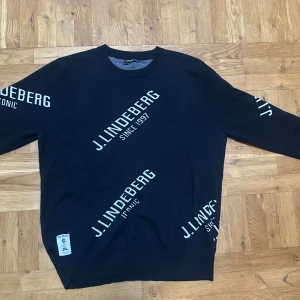Svart J.Lindeberg tröja - Svart sweatshirt från J.Lindeberg i storlek XXL med vit logotyp tryckt flera gånger över hela tröjan. Rund hals och långa ärmar, mjukt material som känns skönt mot huden. Perfekt för dig som gillar streetwear och vill sticka ut med ett coolt print. Köpt för ca 2500kr.