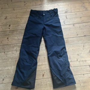 Mörkblå täckbyxor Peak Performance - Säljer ett par mörkblå täckbyxor från Peak Performance med justerbar midja och dragkedjefickor framtill. Byxorna har förstärkta partier vid bensluten och är perfekta för vinterns alla äventyr. Snygg och funktionell design som håller dig varm och torr.