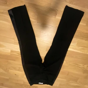 Stylein kostymbyxor  - Säljer ett par svarta kostymbyxor från Stylin med snygga satinband längs sidorna. Byxorna har rak passform.