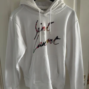 Saint Laurent hoodie - Säljer en vit hoodie från Saint Laurent med färgglatt logotyptryck på bröstet. Tröjan har huva med snörning, känguruficka och långa ärmar. Perfekt för dig som vill ha en clean men ändå unik look.