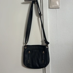 Svart axelväska med blommigt foder - Svart axelväska i skinnimitation med justerbar rem och flera dragkedjefickor både fram och bak. Insidan har ett superfint blommigt tygfoder i beige, rött och blått. Perfekt storlek för mobilen, plånboken och lite till.