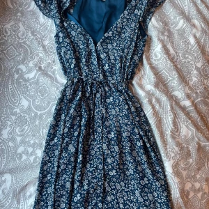 Blommig blå kortklänning från H&M - Supersöt blå kortklänning från H&M med vitt blommönster. Klänningen har omlottliv, resår i midjan och korta ärmar. Perfekt för varma dagar och har en luftig känsla. Materialet är lätt och känns skönt mot huden.