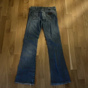 Blå bootcut jeans från Wrangler - Snygga blå jeans från Wrangler med klassisk bootcut-passform. Jeansen har fem fickor, tydliga sömmar och läderpatch med Wrangler-logga bak. Tillverkade i slitstarkt denim med en lätt tvättad look. Perfekta för dig som gillar retrovibbar och avslappnad stil. 