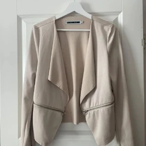 Kavaj - Säljer en stilren beige kavaj från Noisy May i storlek S. Jackan har öppen front, långa ärmar och dekorativa dragkedjor framtill. Perfekt för en trendig och clean look.