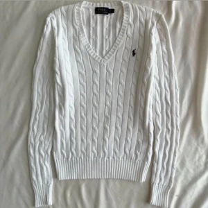 Vit kabelstickad tröja Tommy Hilfiger - Vit kabelstickad tröja från Tommy Hilfiger med v-ringning och långa ärmar. Klassisk design med ribbade muddar och diskret logga på ärmen. Perfekt för dig som gillar en clean och stilren look. Passar mer av en S än M, den sitter dock långt 