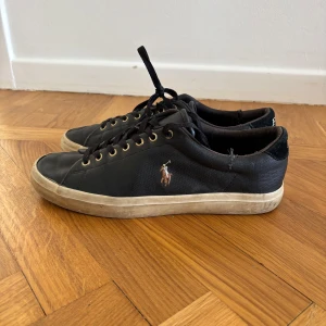 Svarta sneakers från Polo Ralph Lauren - Svarta sneakers i skinn från Polo Ralph Lauren med klassisk logga broderad på sidan och Polo-text bak på hälen. Skorna har svarta snören, guldfärgade öljetter och vit sula. Perfekta för en stilren och avslappnad look.
