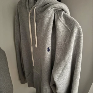 Grå hoodie från Polo Ralph Lauren storlek L - Säljer en klassisk grå hoodie från Polo Ralph Lauren med blå broderad logga på bröstet. Tröjan har huva med vita snören och en stor magficka framtill. Perfekt för chill dagar och enkel att matcha med jeans eller joggers. Skriv för mer information!