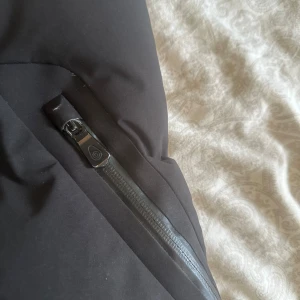 Svart lång dunjacka från Sail racing - Säljer en svart lång dunjacka från Sail racing med huva och dragkedja framtill. Jackan har quiltad design, långa ärmar och praktiska fickor med dragkedja. Perfekt för kalla dagar när du vill hålla dig varm och stilsäker.