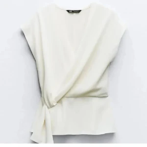 Vit omlottopp från Zara - Stilren vit omlottopp från Zara med knyt i sidan. Toppen har ärmlös design och en snygg v-ringning framtill. Perfekt för dig som gillar minimalistisk och clean stil. Lätt och skön att bära, passar till många olika outfits.
