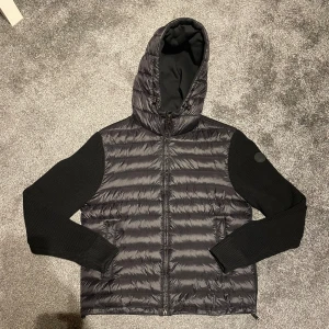 Svart Colmar cardigan med huva - Snygg svart cardigan från Colmar med stickade ärmar och vadderad framsida. Jackan har huva, dragkedja och diskret logga på ärmen. Perfekt för dig som vill ha en modern look med mix av material och detaljer. Digitalt kvitto finns 👍det finns inga tecken på användning har bara prövat den, beställde en storlek mindre men fick denna, var för snabb på att ta bort lapparna men har som sagt digitalt kvitto på den 🙂