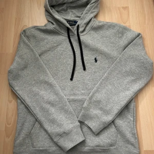 Grå Polo Ralph Lauren hoodie - Snygg grå hoodie från Polo Ralph Lauren med svart snörning och klassisk broderad logga på bröstet. Tröjan har huva, känguruficka och är tillverkad i mjuk bomullsmix. Perfekt för chill dagar eller när du vill ha en clean look. Passar perfekt året om! Sälj pga har flera st! Pris kan diskuteras!!! S passar även M