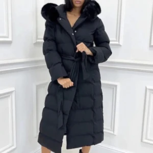 Svart lång dunjacka med fuskpäls - Säljer en svart lång dunjacka med stor huva och fuskpälsdetaljer. Jackan har bälte i midjan, knappar framtill och är quiltad för extra värme. Perfekt för kalla vinterdagar och ger en riktigt snygg siluett.