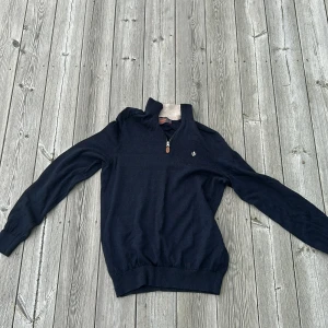 Marinblå merinoulltröja från Morris - Snygg marinblå långärmad tröja från Morris i 100% merinoull. Tröjan har half zip med liten läderdetalj och ribbade muddar. Perfekt för dig som gillar klassisk stil med en modern touch. Har tyvärr en defekt vid vänster armbåge men är annars i bra skick!