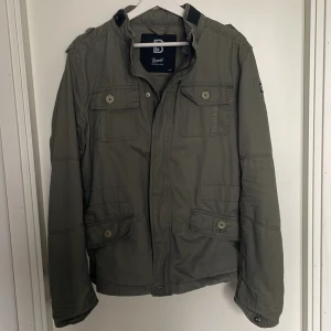 Grön FieldJacket - Säljer en olivgrön Fieldjacket från Brandit med flera fickor framtill, axelremmar och snygga knappar. Jackan har dragkedja och knäppning, samt en patch med Brandit-loggan på ärmen. Perfekt för dig som gillar en stilren och funktionell look, Pris kan diskuteras! 