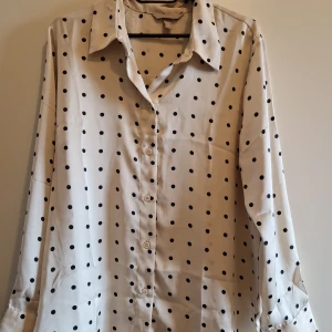 Prickig vit skjorta från H&M, M - Snygg vit skjorta från H&M i storlek M med svarta prickar. Klassisk krage, knäppning framtill och långa ärmar. Skjortan har en rak passform och är tillverkad i ett mjukt, lätt material som känns skönt mot huden. Perfekt för dig som gillar en stilren men lekfull look.