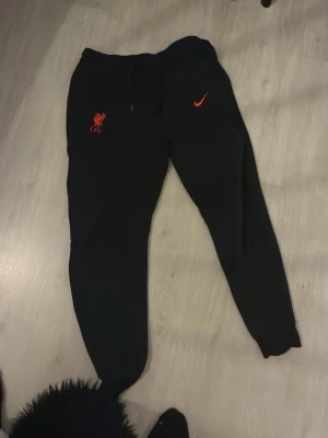 Svarta Nike Liverpool mjukisbyxor M - Svarta mjukisbyxor från Nike med Liverpool FC-logga i rött och röd swoosh på framsidan. Byxorna har snörning i midjan och ribbade muddar vid bensluten. Perfekta för chill eller sport, med skön passform och mjukt material.