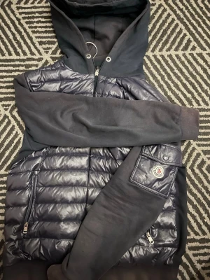 Svart pufferjacka med huva från Moncler - Svart pufferjacka från Moncler med huva och quiltad framsida i glansigt material. Jackan har dragkedjor på fickorna och en snygg Moncler-logga på ärmen. Ärmar och rygg är i mjuk bomull, vilket ger en cool kontrast till den blanka fronten. Har hål 🕳️ i både fickorna 
