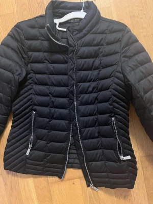 Svart quiltad pufferjacka från Amisu - Svart pufferjacka från Amisu med quiltad design och snygga dragkedjor på fickorna. Jackan har en hög krage och är tillverkad i polyester, perfekt för kyliga dagar. Passformen är normal och jackan har långa ärmar.