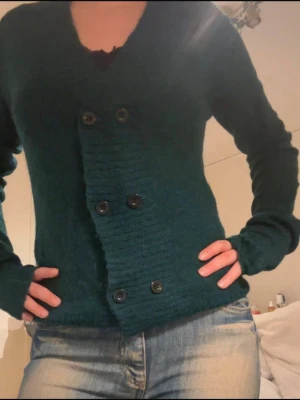 Mörkgrön stickad kofta med knappar - Snygg mörkgrön stickad kofta med dubbelknäppning framtill och ribbade detaljer. Koftan har lång ärm och v-ringad hals, perfekt att slänga på över en t-shirt eller linne. Passformen är normal och materialet känns mjukt och skönt mot huden.