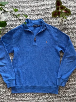 Blå half zip tröja från Polo Ralph Lauren - Säljer en blå långärmad tröja från Polo Ralph Lauren med half zip och klassisk broderad logga på bröstet. Tröjan är stickad i bomull och har ribbade muddar vid ärmslut och nederkant. Perfekt för dig som gillar stilren och sportig look. Nytt skick. 