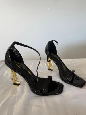 Svarta Saint Laurent klackar - Säljer ett par svarta klackar från Saint Laurent med glansig finish och ikonisk guld YSL-klack. Skorna har smal rem över tårna, fyrkantig tå och justerbar axelrem. Helt oanvända. Storlek 38