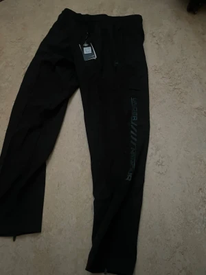 Svarta träningsbyxor från Under Armour - Svarta träningsbyxor från Under Armour med reflekterande logga och text längs benet. Byxorna har snörning i midjan och dragkedja vid benslut. Materialet är lätt och syntetiskt, perfekt för sport och aktivitet. Snygg, loose passform med modern look.