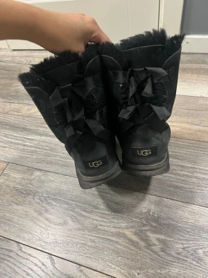 Svarta UGG boots med rosetter - Mysiga svarta UGG boots i mocka med fluffigt foder och dekorativa rosetter bak. Klassisk rund tå och platt sula för maximal komfort. Perfekta för kalla dagar och ger en chill vibe till din outfit.