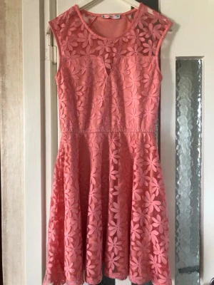 Rosa blommig kortklänning - Supersöt rosa kortklänning med stora blommor i spets över hela klänningen. Ärmlös modell med rund halsringning och lätt markerad midja. Kjolen är utställd och tyget känns lätt och luftigt, perfekt för varma dagar.