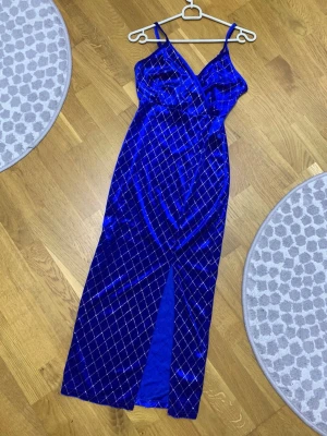 Blå glittrig klänning i sammet  - Supersnygg blå långklänning i mjuk sammet med tunna axelband och omlott-v-ringning. Klänningen har ett rutigt mönster med glittriga detaljer och en slits framtill som ger extra edge. Perfekt för dig som vill sticka ut!