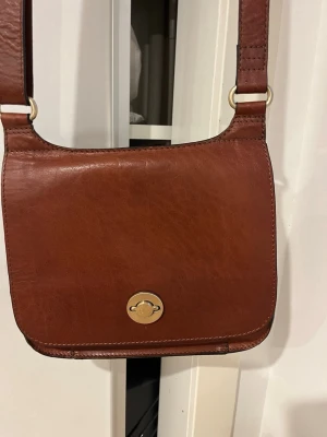 Brun axelväska i skinn från Saddler - Snygg brun axelväska i skinn från Saddler med justerbar rem och guldfärgat spänne framtill. Väskan har ett klassiskt, stilrent utseende och ett innerfack med dragkedja. Perfekt för dig som vill ha en tidlös och praktisk väska. 15x15cm