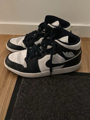 Nike Air Jordan 1 Mid mörk blå/Vit 43 - Säljer ett par Nike Air Jordan 1 Mid i mörkblå och vitt. Skorna har klassisk snörning, rund tå och perforerat skinn på ovandelen. Snygg kontrast mellan svart och vit, med ikonisk Air Jordan-logga på sidan. Perfekt för dig som gillar streetwear och sneakers med retro vibe.