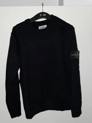 Svart tröja från Stone Island - Svart tröja från Stone Island i storlek L. Klassisk rund hals och långärmad modell med den ikoniska logotypen på vänster ärm. Tröjan har ribbade muddar vid hals, ärmslut och nederkant. Perfekt för dig som gillar stilrena och tidlösa plagg.
