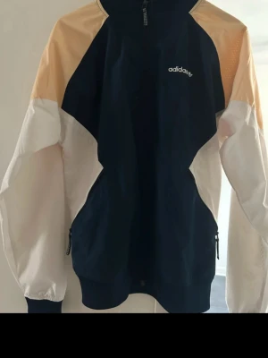 Adidas vindjacka i blått och beige - Snygg vindjacka från Adidas med färgblock i marinblått, beige och vitt. Jackan har dragkedja framtill, höga muddar och två sidofickor med dragkedja. Perfekt för chill dagar och sportiga outfits.