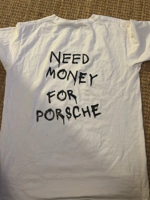 Vit t-shirt med tryck från Bvaldi - Vit t-shirt från Bvaldi i bomull med svart tryck på ryggen där det står 'Need money for Porsche' i en cool, handskriven stil. Framtill finns en liten svart logga på bröstet. Klassisk rund hals och korta ärmar, perfekt för dig som gillar statement-plagg.