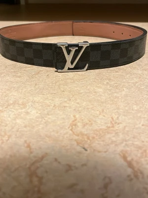 Louis Vuitton rutig svart skinnbälte - Säljer ett Louis Vuitton bälte med klassiskt LV-spänne i silver. Bältet är svart med rutigt mönster och har en insida i ljusbrunt skinn. Den är använd men fortfarande mycket användbart och praktiskt.