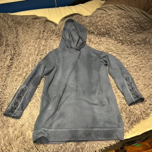 Blå lång hoodie med snörning - Blå lång hoodie med huva och snörningsdetaljer på båda ärmarna.  Hoodien har fickor på sidorna och är gjord i mjukt bomullsmaterial.  Perfekt för chill dagar och har en loose passform som gör den extra bekväm. Den är i använt skick. Storlek L. Från HOLLISTER.
