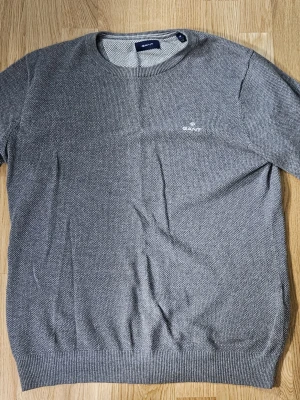 Grå stickad tröja från GANT - Snygg grå stickad tröja från GANT i storlek M. Tröjan har rund hals, diskret logga på bröstet och ribbade muddar vid ärmslut och nederkant. Perfekt för dig som gillar klassisk och enkel stil med en modern touch. Kan också användas över en skjorta för en stilren look.