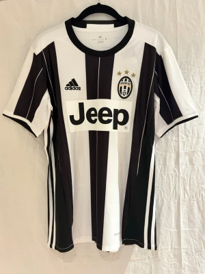 Juventus tröja Dybala 16/17 - Juventus matchtröja med Dybala 21 på ryggen. Klassisk svartvit randig design, broderat klubbmärke med tre guldstjärnor och Jeep-logga på bröstet. Tillverkad i lätt polyester med climacool-teknologi för bästa komfort under match. 