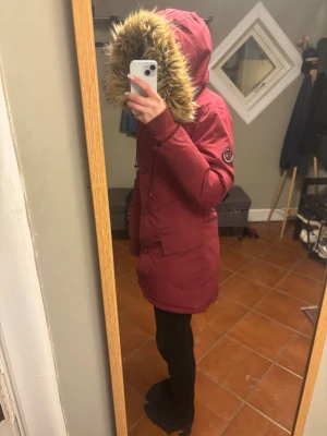 Mörkröd jacka med fuskpäls - Säljer en mörkröd jacka med huva och fuskpäls. Jackan har snörning i midjan, stora fickor med lock och knappar framtill. Perfekt för kalla dagar och har en normal passform. Tyvärr är dragkedjan sönder. 